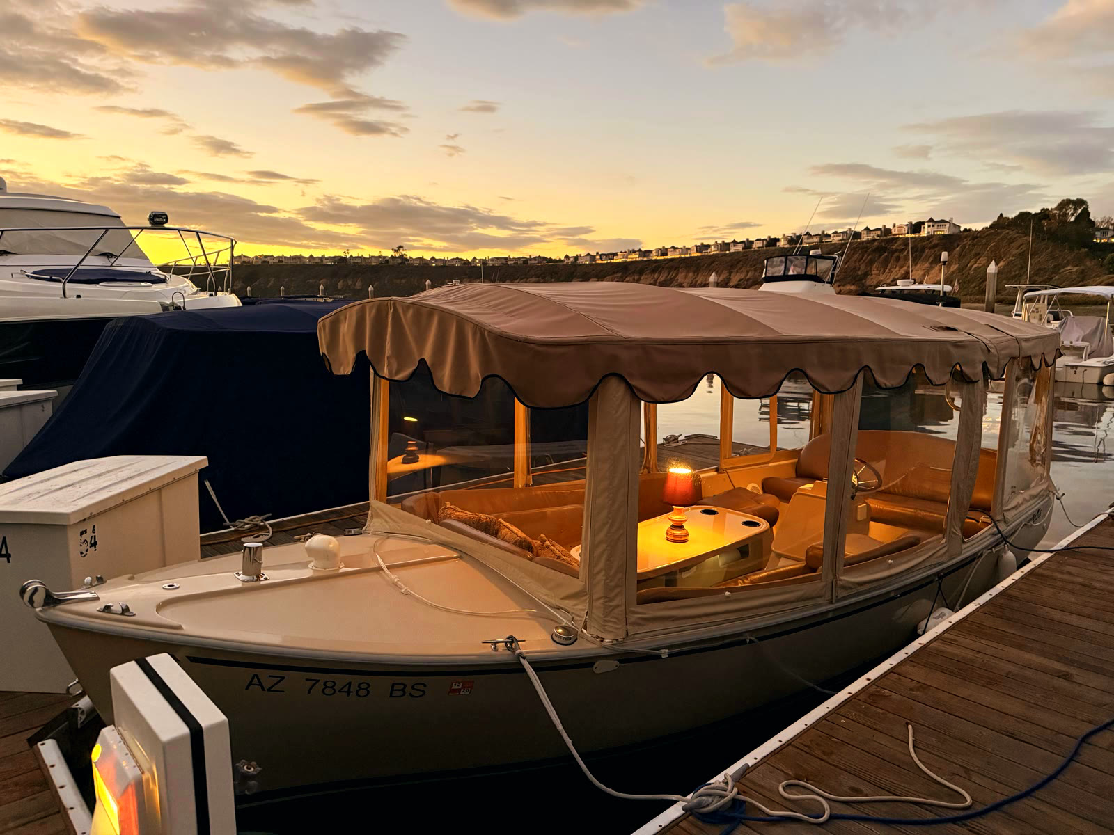 A Duffy electric boat with a warm cabin lamp at its Bayside Village slip at dusk, with the last of the sunset over the Back Bay bluffs behind