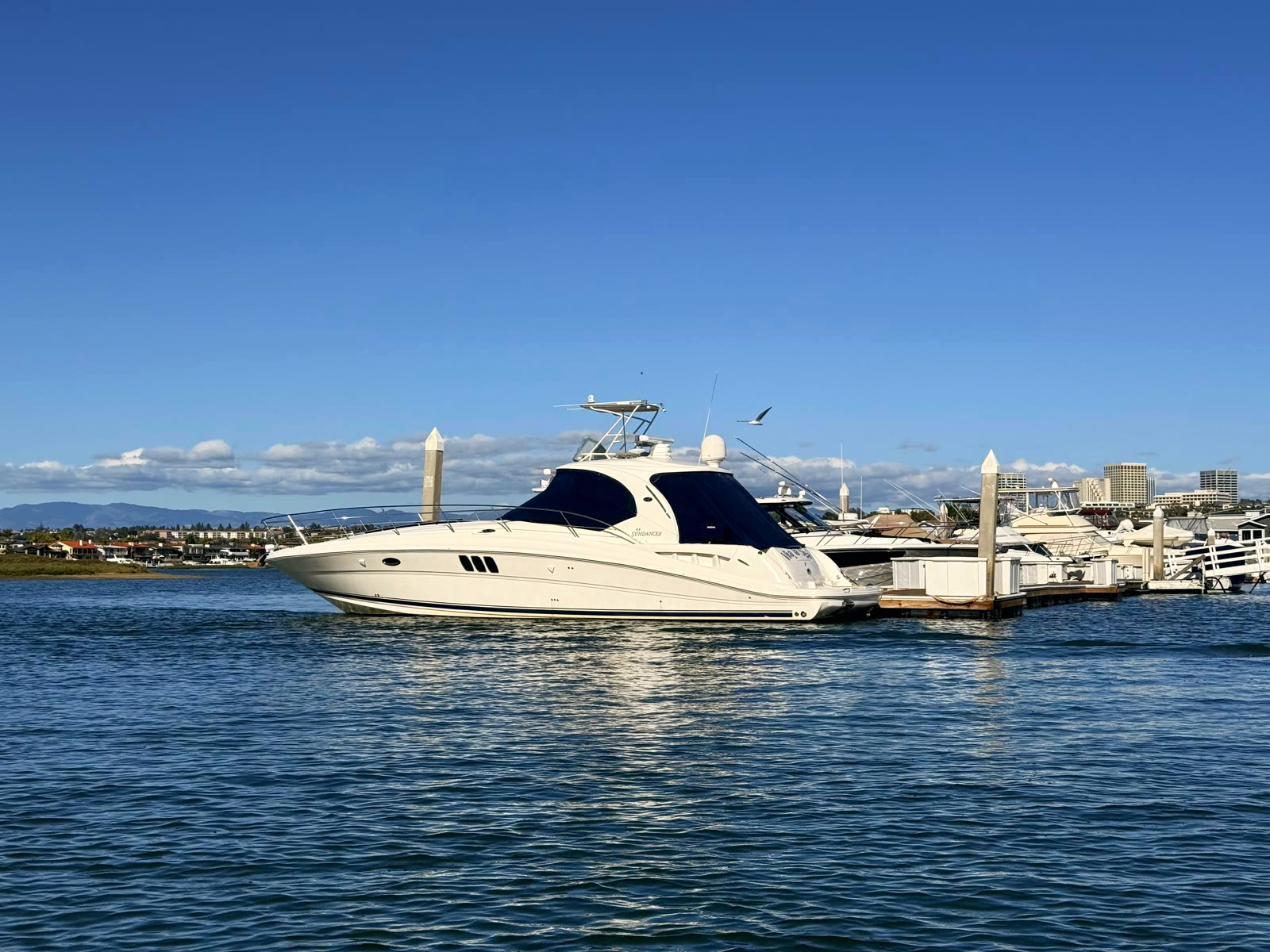 A Sea Ray Sundancer at her Bayside Village Marina end tie on calm water, with the Newport Center skyline on the horizon