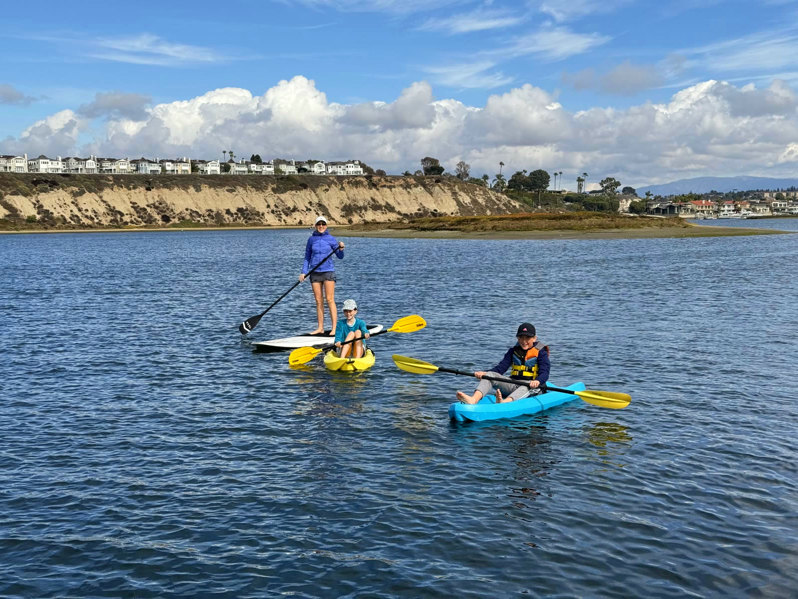 A family paddleboarding and kayaking on calm Back Bay water off the Bayside Village docks, with the Castaways bluff and Dover Shores homes behind
