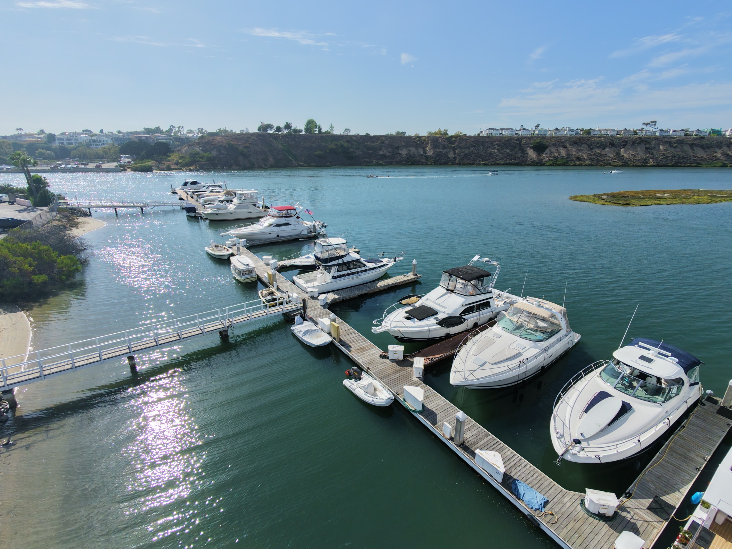 Aerial view of the Bayside Village dock fingers on calm water, with the marsh islands of Upper Newport Bay and the Castaways bluff across the channel