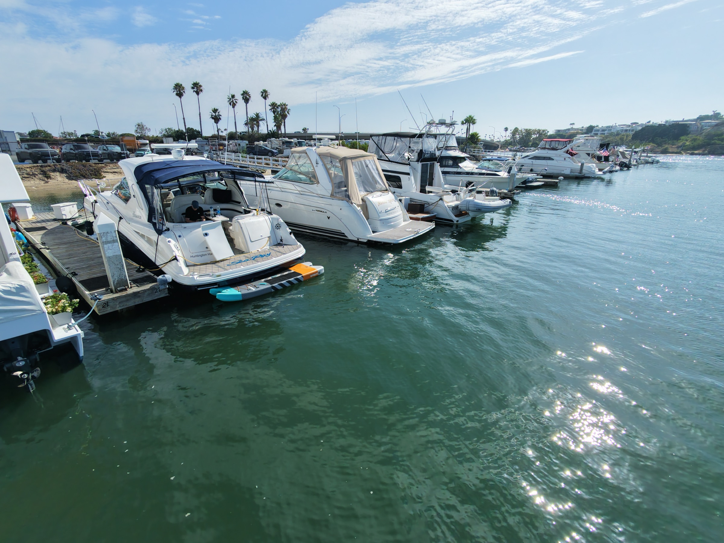 Dock-level view of cruisers in their Bayside Village slips on sparkling water, with palms and the PCH Bay Bridge behind