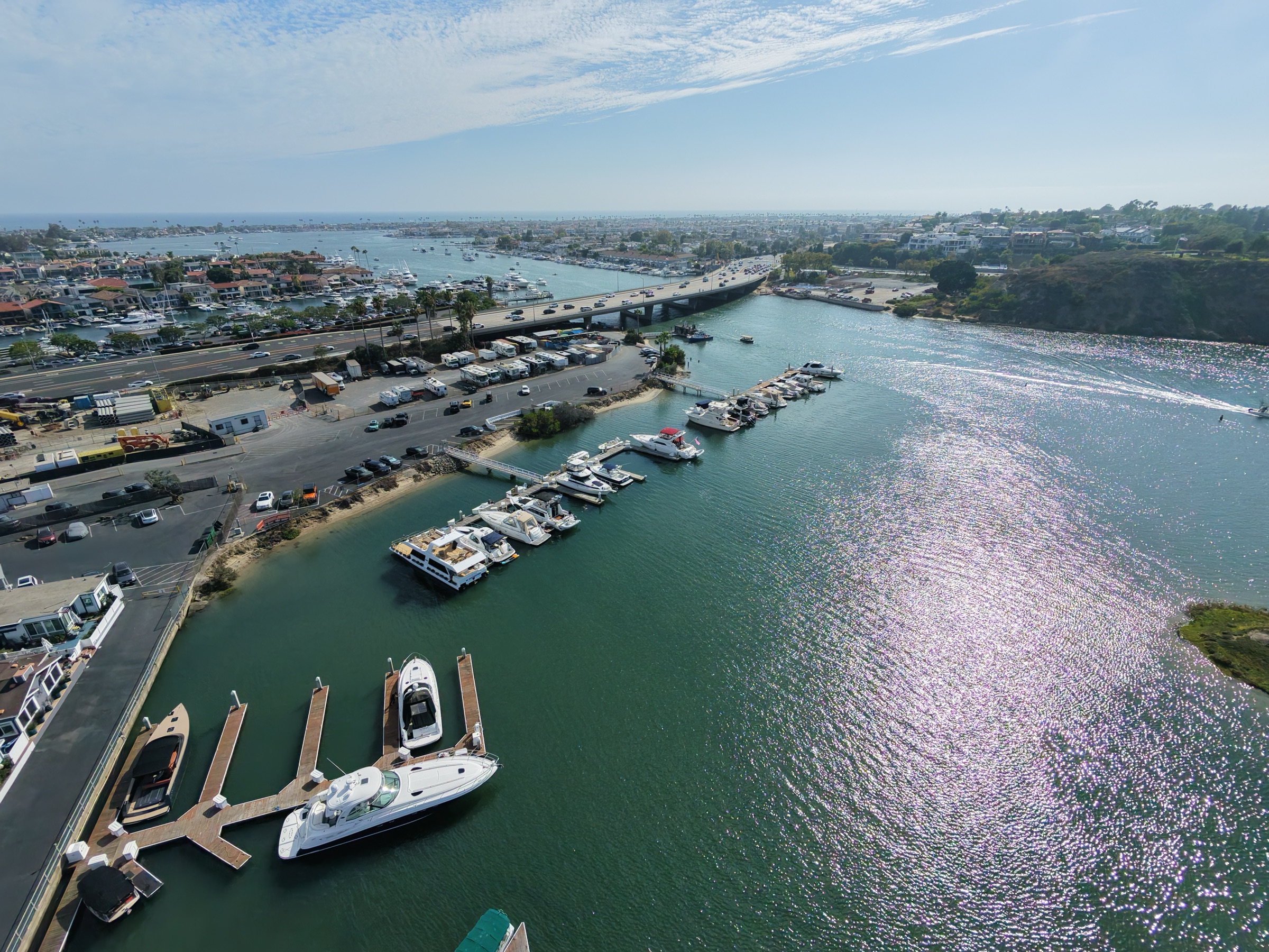 Aerial view down the sparkling channel from the Bayside Village docks toward the PCH Bay Bridge and Lower Newport Harbor, with the ocean on the horizon