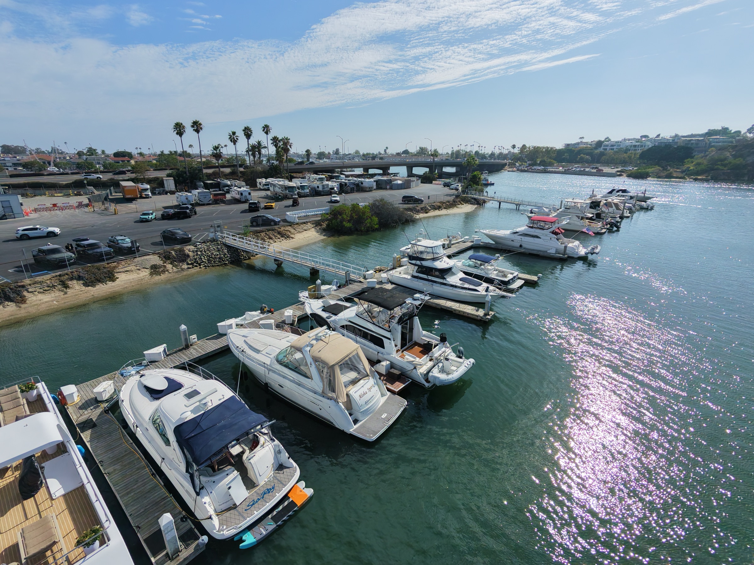 Aerial view of boats in their slips at the Bayside Village docks, with the gangway, palms, and the PCH Bay Bridge behind