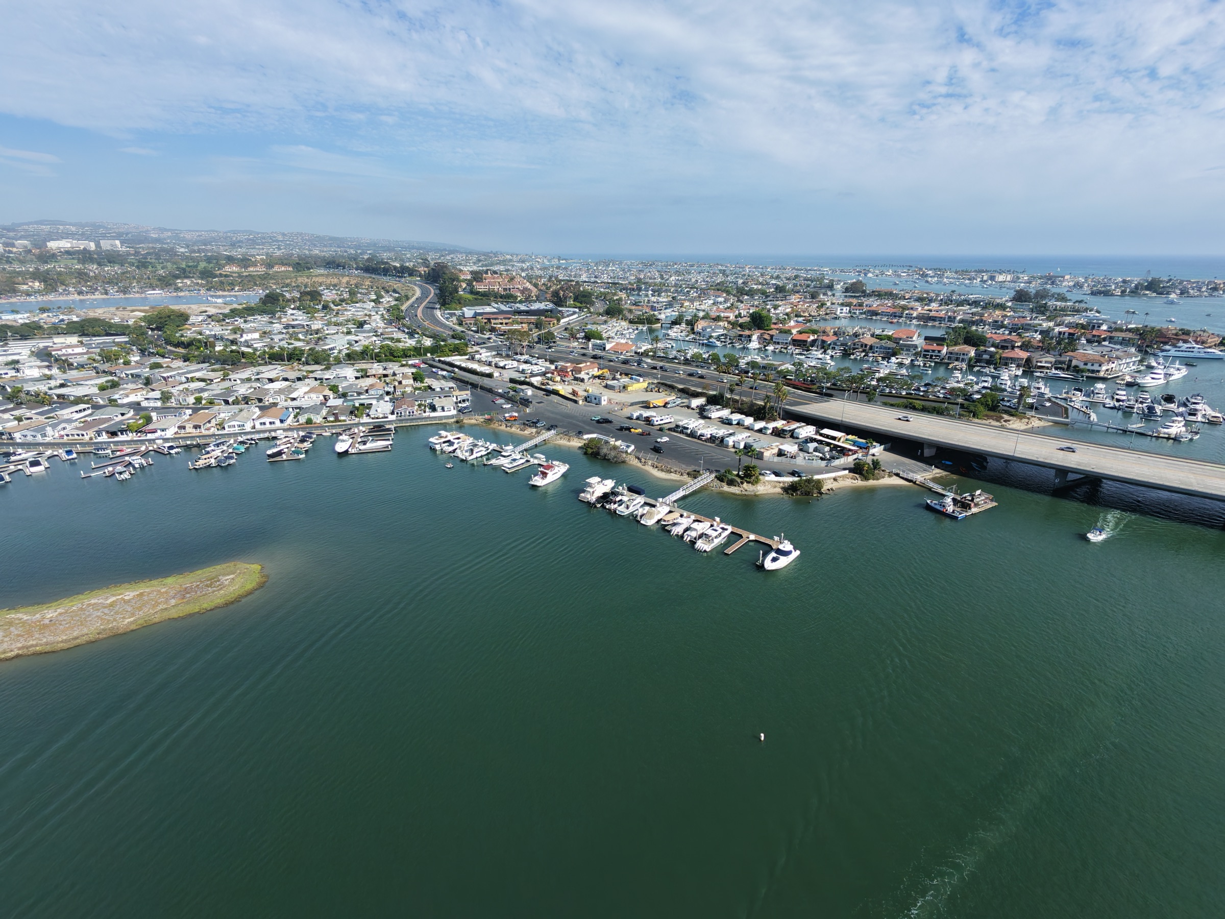 Wide aerial of the seam where the PCH Bay Bridge divides Newport Harbor — the Bayside Village docks in the foreground, Lower Newport Harbor and the coast beyond