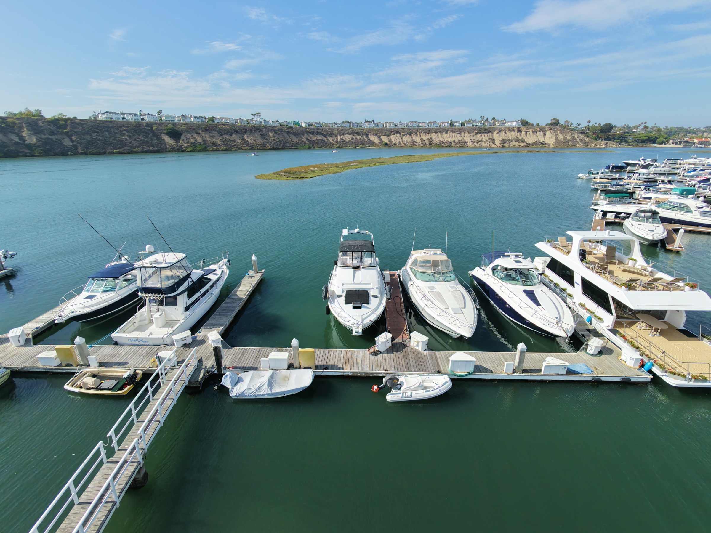 Boats bow-in at the Bayside Village dock with the marsh island of Upper Newport Bay and the Dover Shores bluff across the calm channel