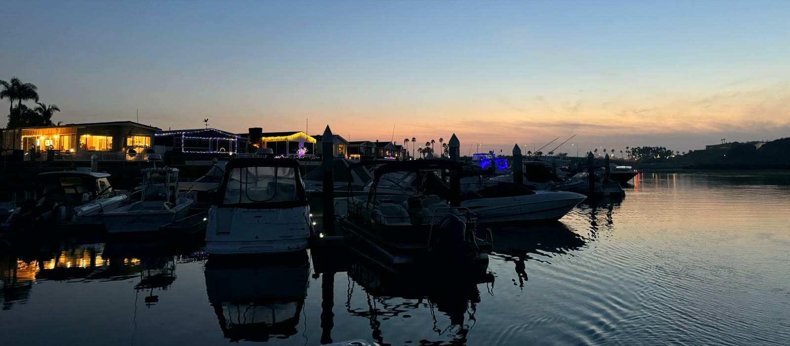 Dusk at Bayside Village Marina — village homes glowing with string lights behind boats in their slips, the calm Back Bay channel reflecting the last of the sunset