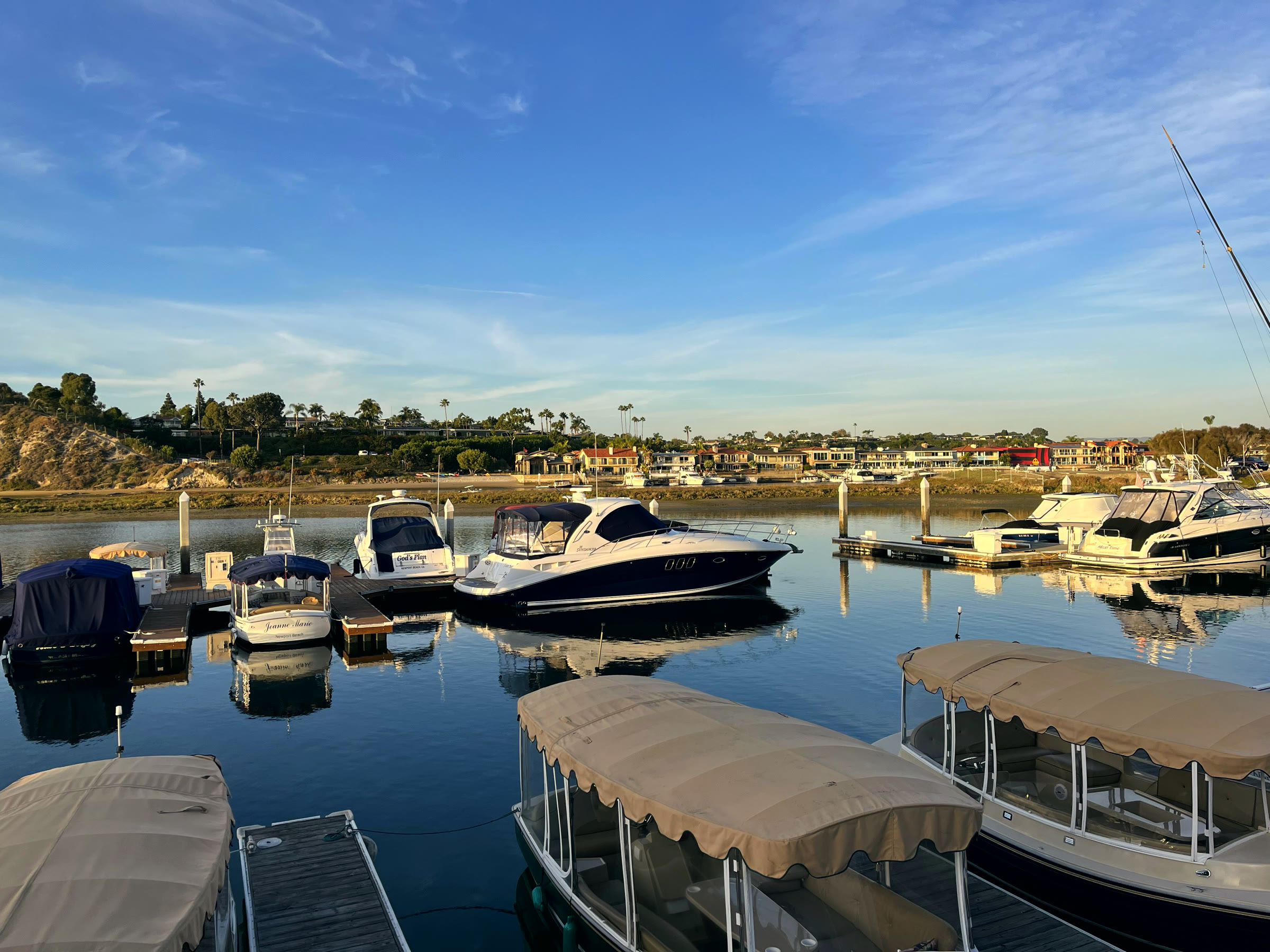 Morning at Bayside Village Marina — Duffy boats under canvas tops and Sea Ray cruisers at their slips on glassy water, with the Back Bay bluffs and Corona del Mar homes across the channel