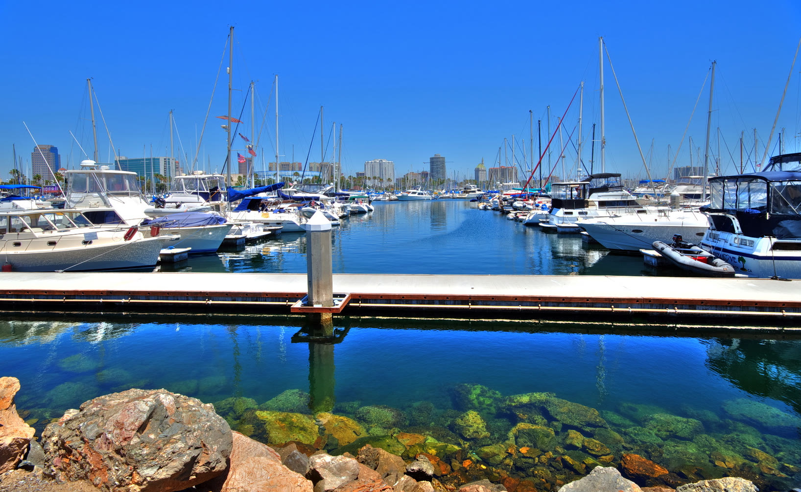 Sailboats and powerboats at their docks in Rainbow Harbor on calm Queensway Bay, with the downtown Long Beach skyline rising behind the masts
