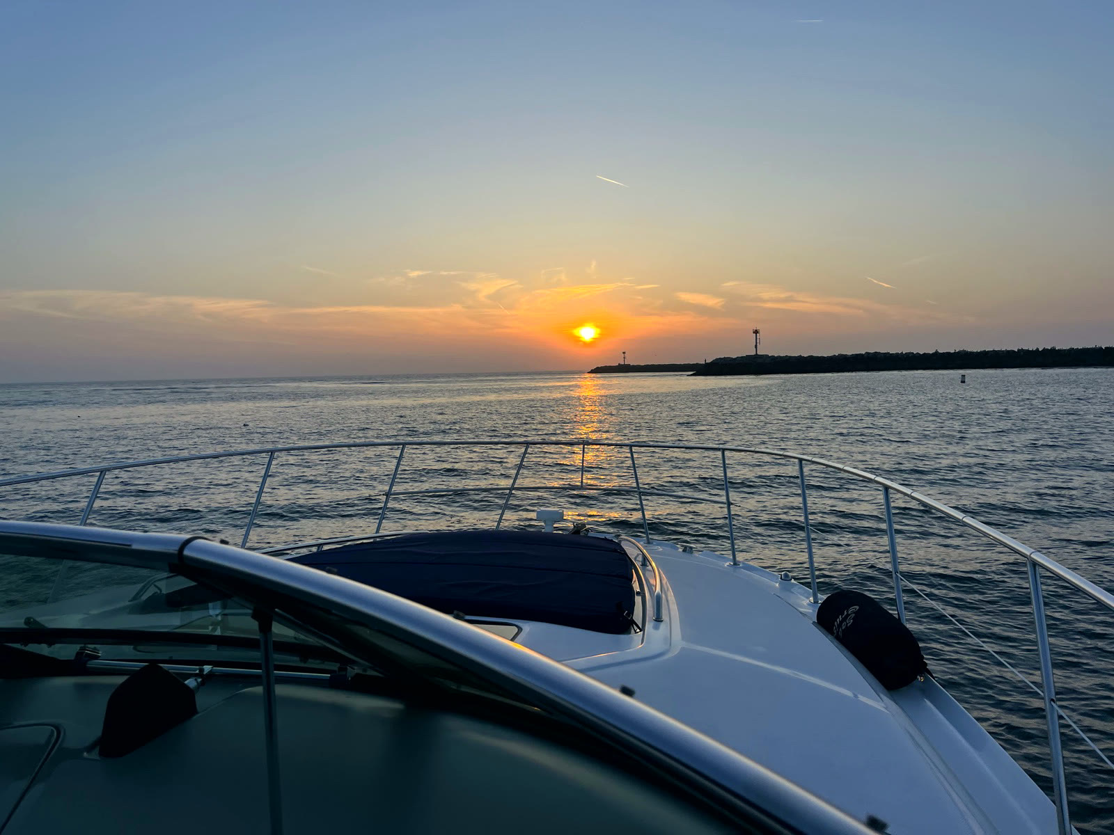 View over a boat's bow rail toward the Newport Harbor jetty entrance at sunset, with the sun low over calm water and the rock breakwater silhouetted