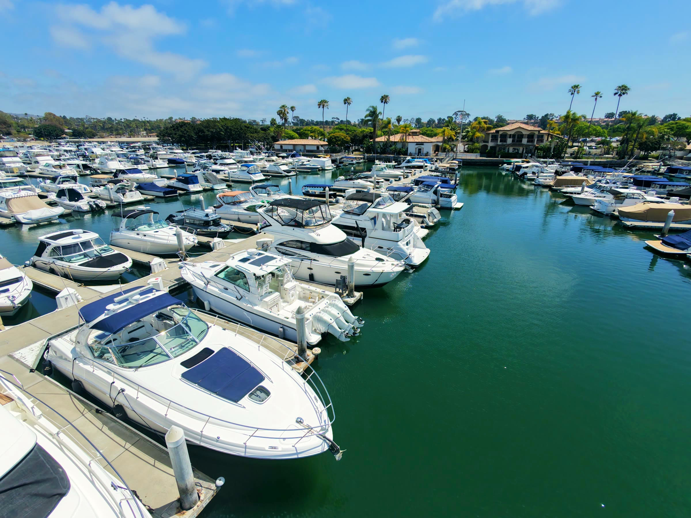 Low aerial of the Newport Dunes Marina basin — rows of powerboats in single-loaded slips, palms and the marina buildings along the shore