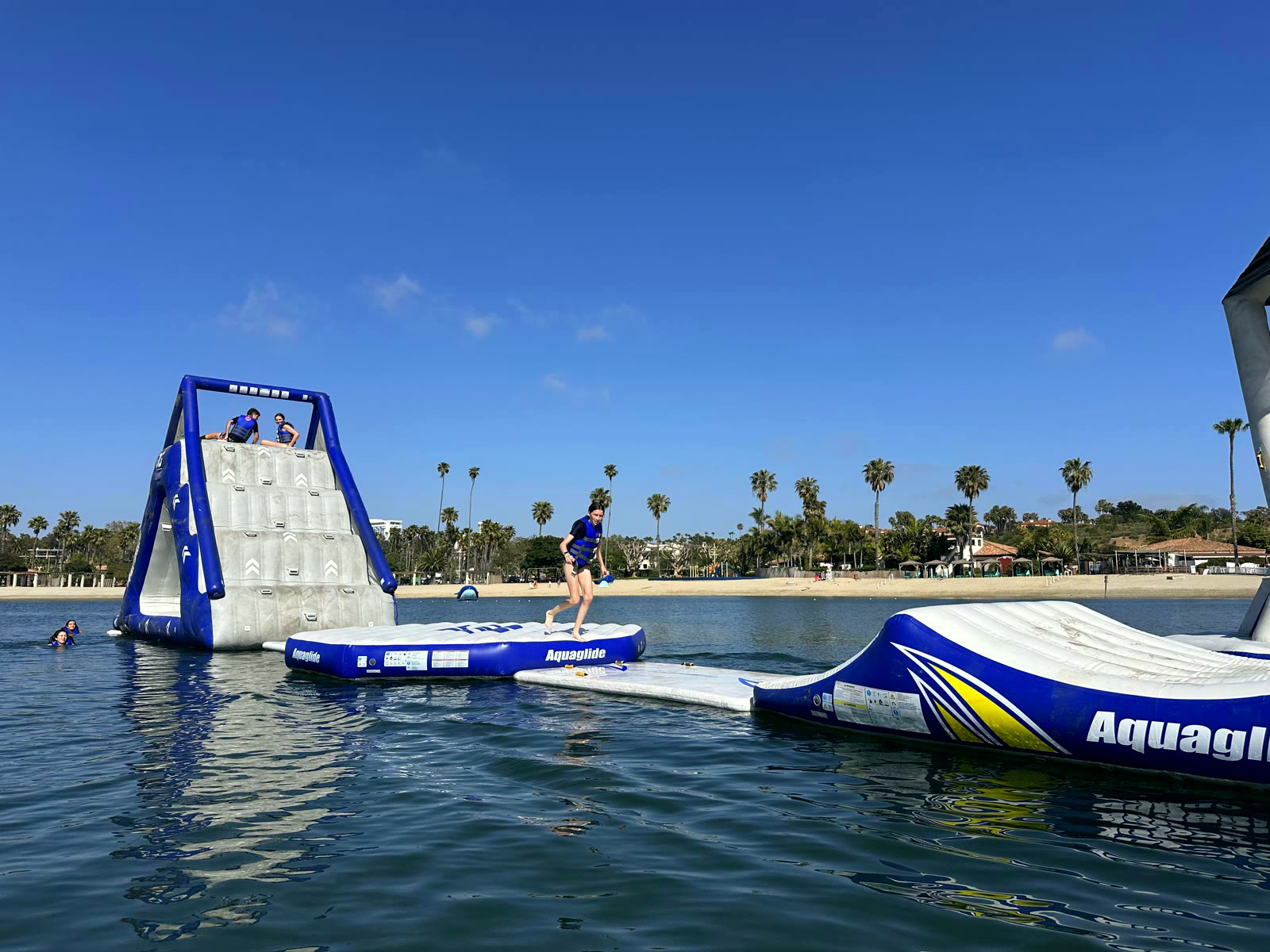 Kids climbing an inflatable water-park tower on the Newport Dunes lagoon, with the beach and palm rows behind