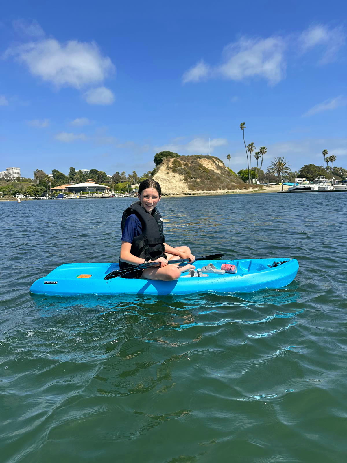 A kid in a life jacket paddling a kayak on the calm Back Bay off Newport Dunes, bluffs and moored catamarans behind