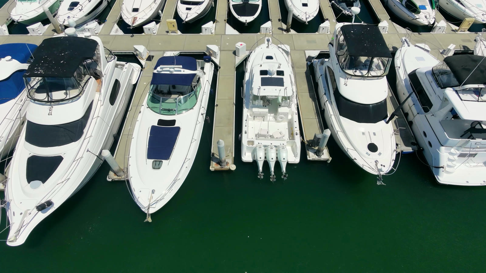 Straight-down aerial of powerboats in their slips at Newport Dunes Marina, white hulls and canvas covers on green Back Bay water