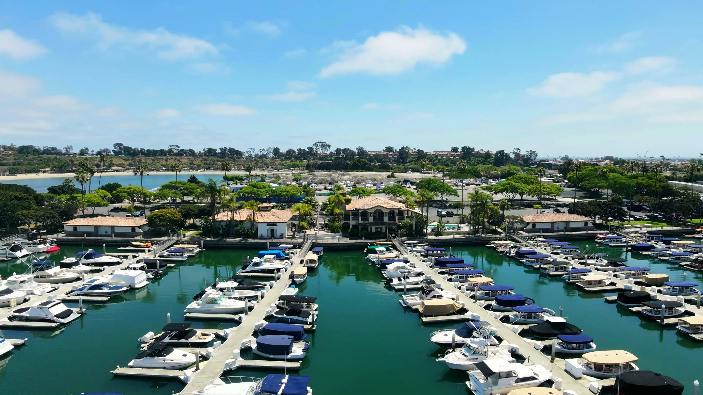 Aerial view of Newport Dunes Marina — rows of boats in single-loaded slips in the protected basin, the marina building at the head of the docks, and the lagoon beach and resort grounds beyond